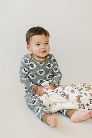 A toddler wearing a green Aztec-patterned pajama set holds a patterned blanket, sitting on a light background.