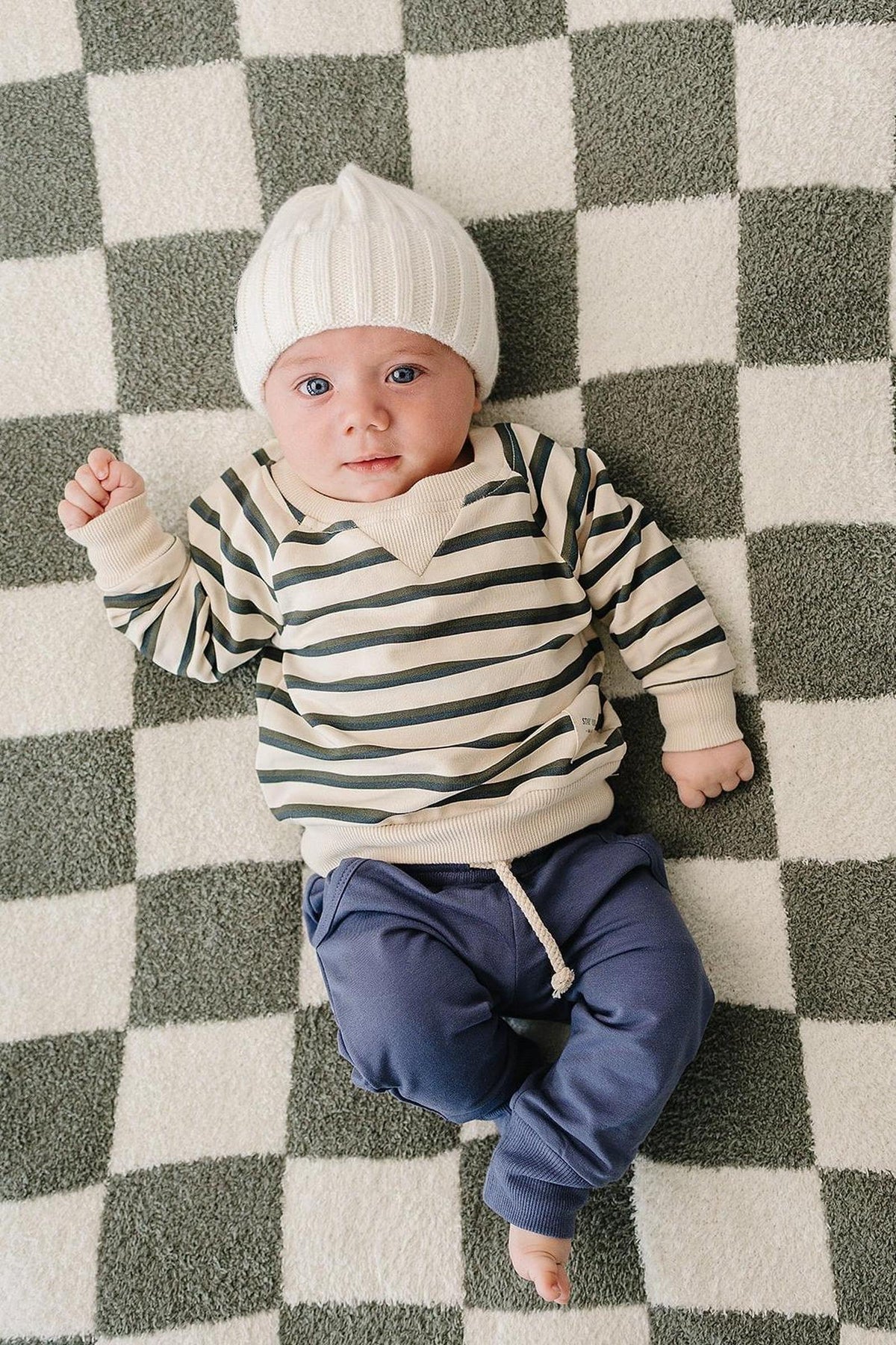 A baby lies on a checkered blanket, wearing a striped long-sleeve crewneck shirt and blue pants, topped with a white knitted beanie.