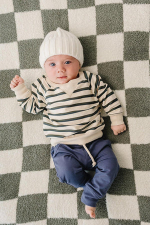 A baby lies on a checkered blanket, wearing a striped long-sleeve crewneck shirt and blue pants, topped with a white knitted beanie.