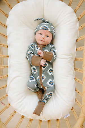 A baby in a basket wears green Aztec-patterned pajamas and matching hat, lying on a white cushion.