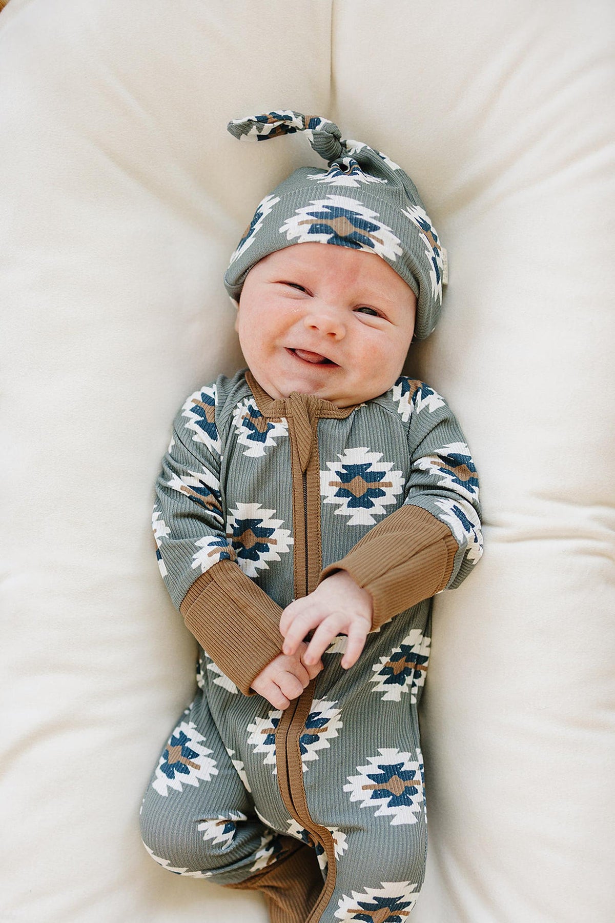 Smiling baby in green Aztec-patterned bamboo pajamas and matching hat, lying on a soft, white cushion.