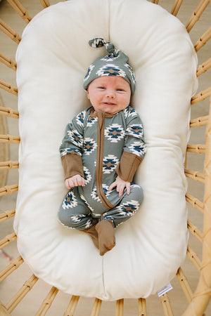 A baby wearing a green Aztec-patterned bamboo pajama and hat lies on a white cushion in a wicker bassinet.
