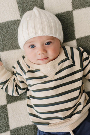 A baby lies on a checkered blanket, wearing a cream and green striped long-sleeve crewneck shirt and a white knit hat.