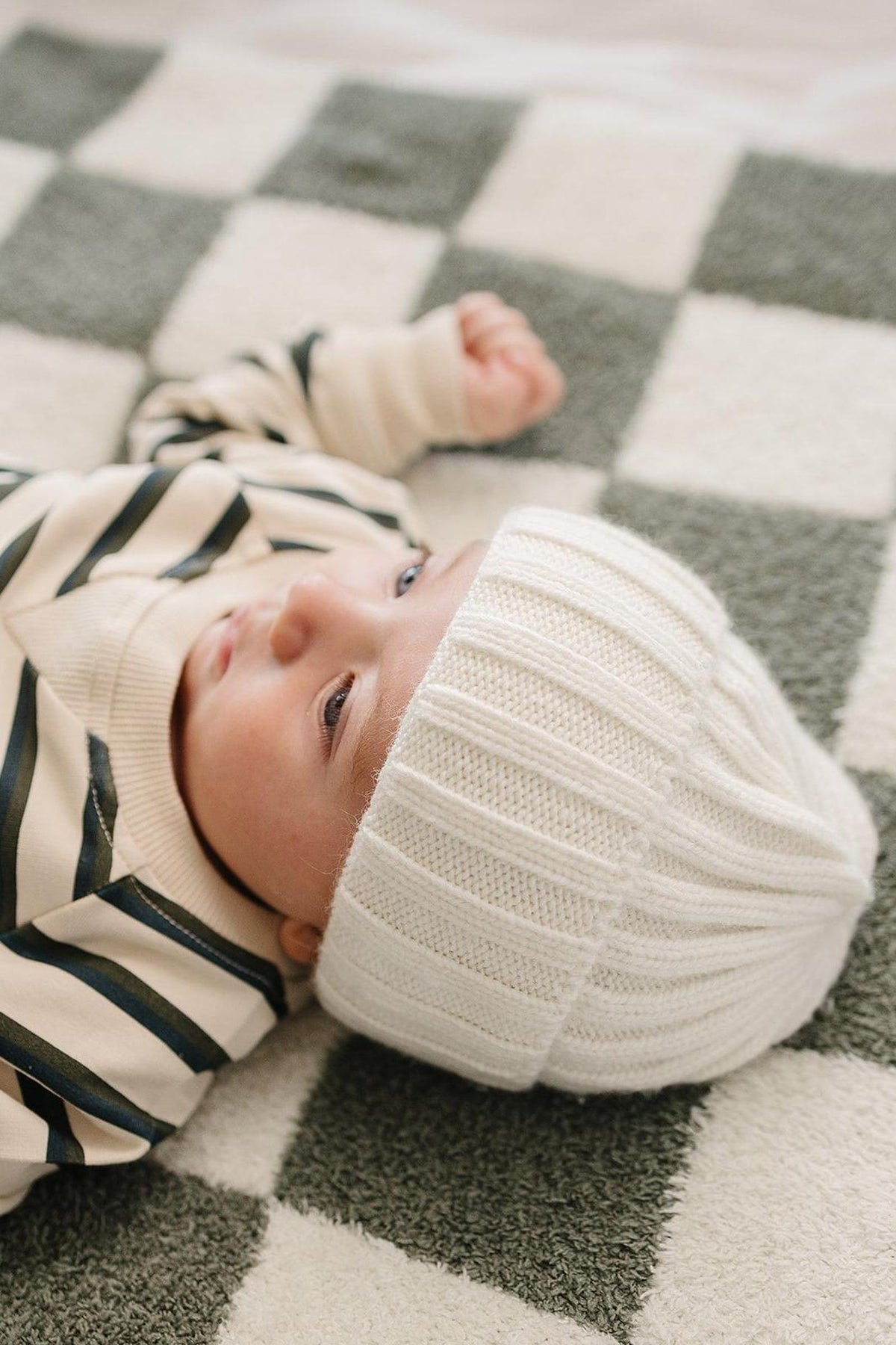 A baby wearing a white chunky knit beanie lies on a checkered blanket, looking up with curious eyes.