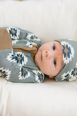 A baby wears green Aztec-patterned pajamas with a matching hat, lying on a white cushion.
