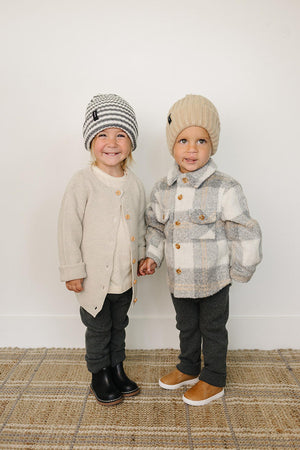 Two children wearing cozy knit beanies and warm sweaters, standing on a woven rug, smiling warmly.