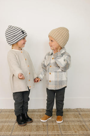 Two young children hold hands, wearing cozy knit beanies and stylish outfits, against a neutral background with a textured rug.
