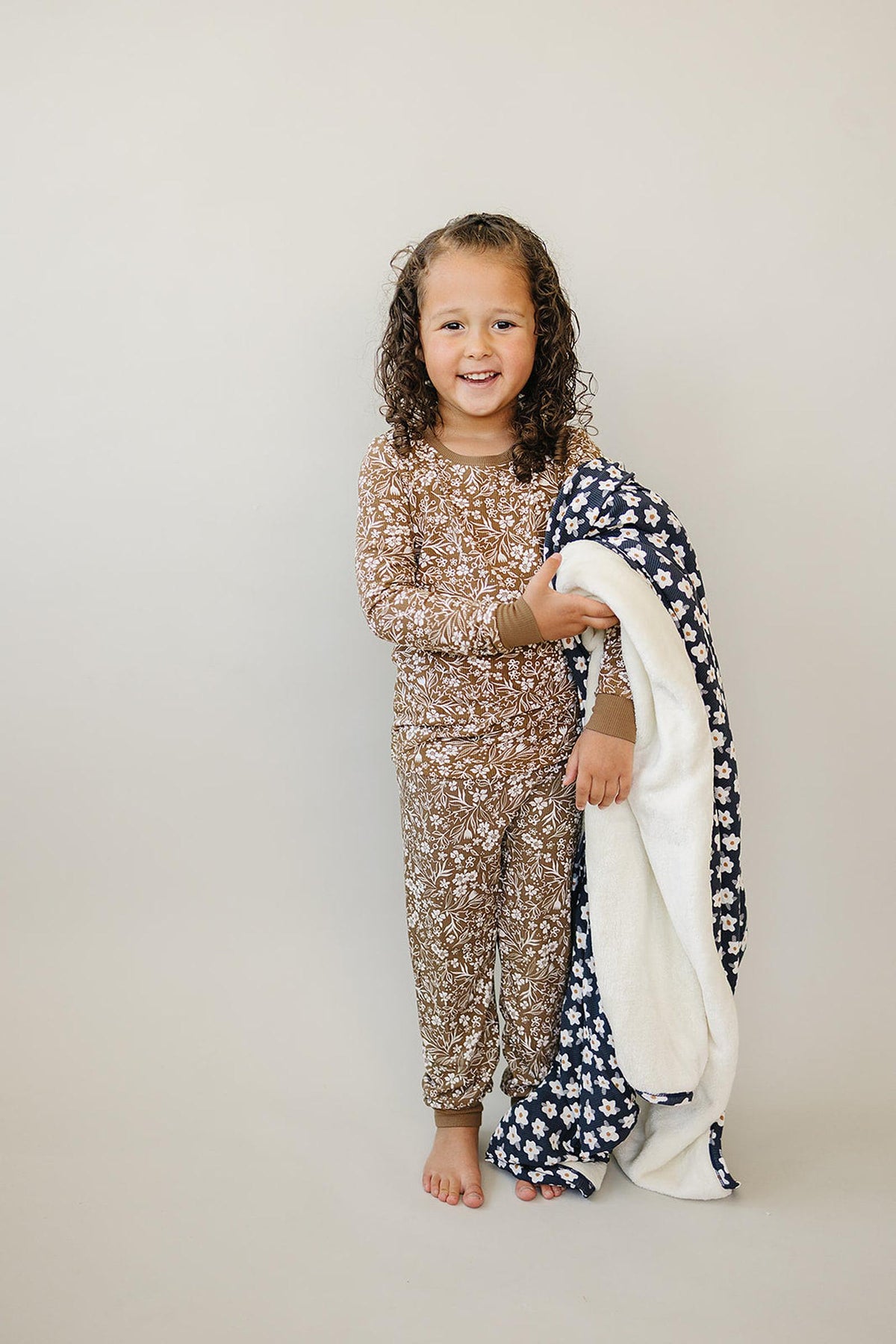 Child in brown floral pajama set holding a navy floral blanket, standing against a plain background.