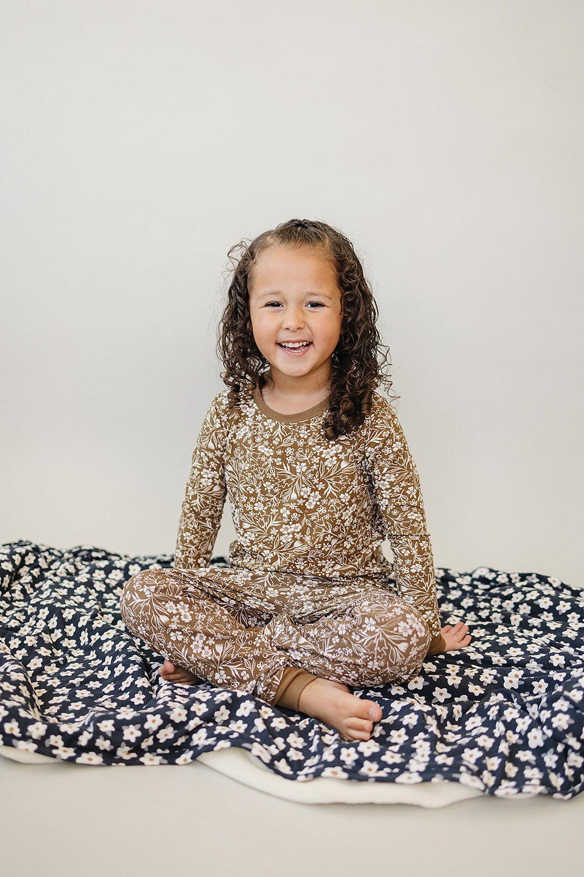A child in a brown floral pajama set sits on a blue floral blanket, smiling at the camera.