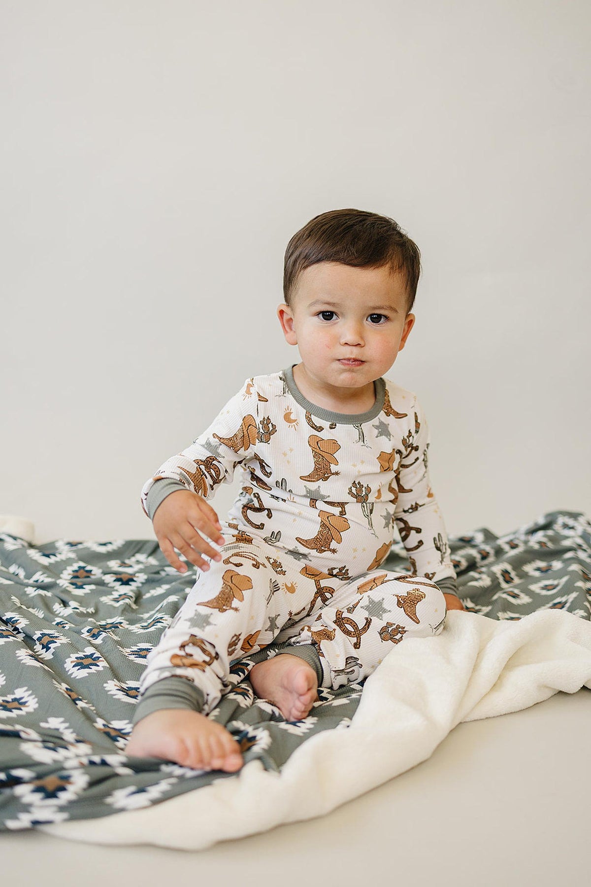 A toddler wearing a cowboy-themed pajama sits on a patterned blanket, featuring a soft, neutral backdrop.