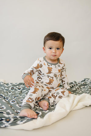 A toddler wearing a cowboy-themed pajama sits on a patterned blanket, featuring a soft, neutral backdrop.