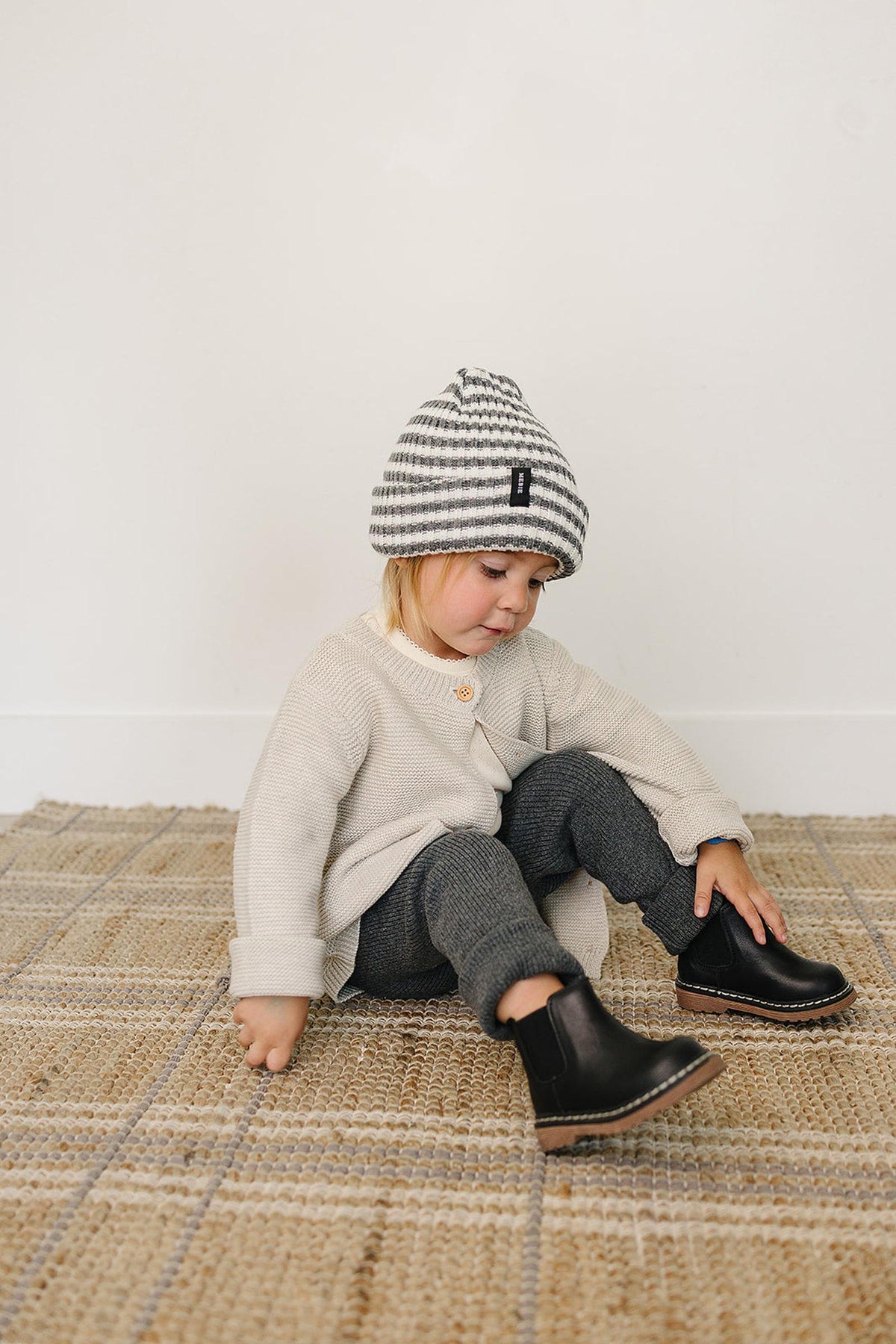A toddler sits on a woven rug, wearing a grey and white striped beanie, beige sweater, and black boots.