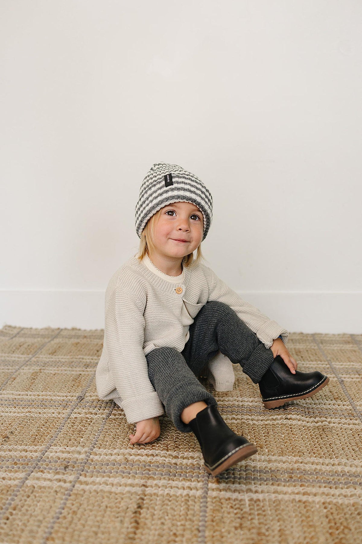 A child sits on a woven rug, wearing a grey and white striped beanie, cream sweater, and black boots.