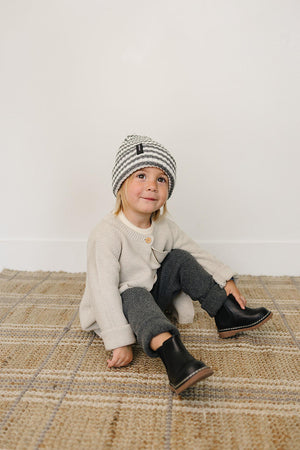 A child sits on a woven rug, wearing a grey and white striped beanie, cream sweater, and black boots.