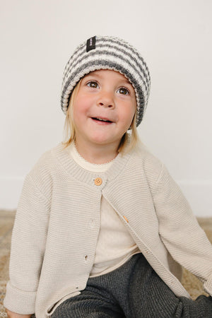 A cheerful child wearing a grey and white striped chunky knit beanie, styled with a cozy beige cardigan and gray.