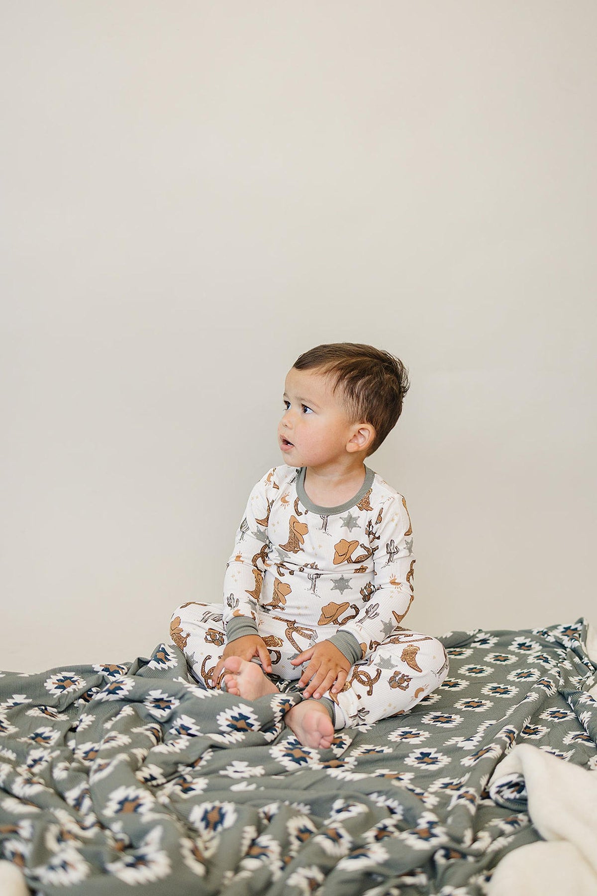 A toddler sits on a patterned blanket wearing a white cowboy-themed pajama set with green cuffs.