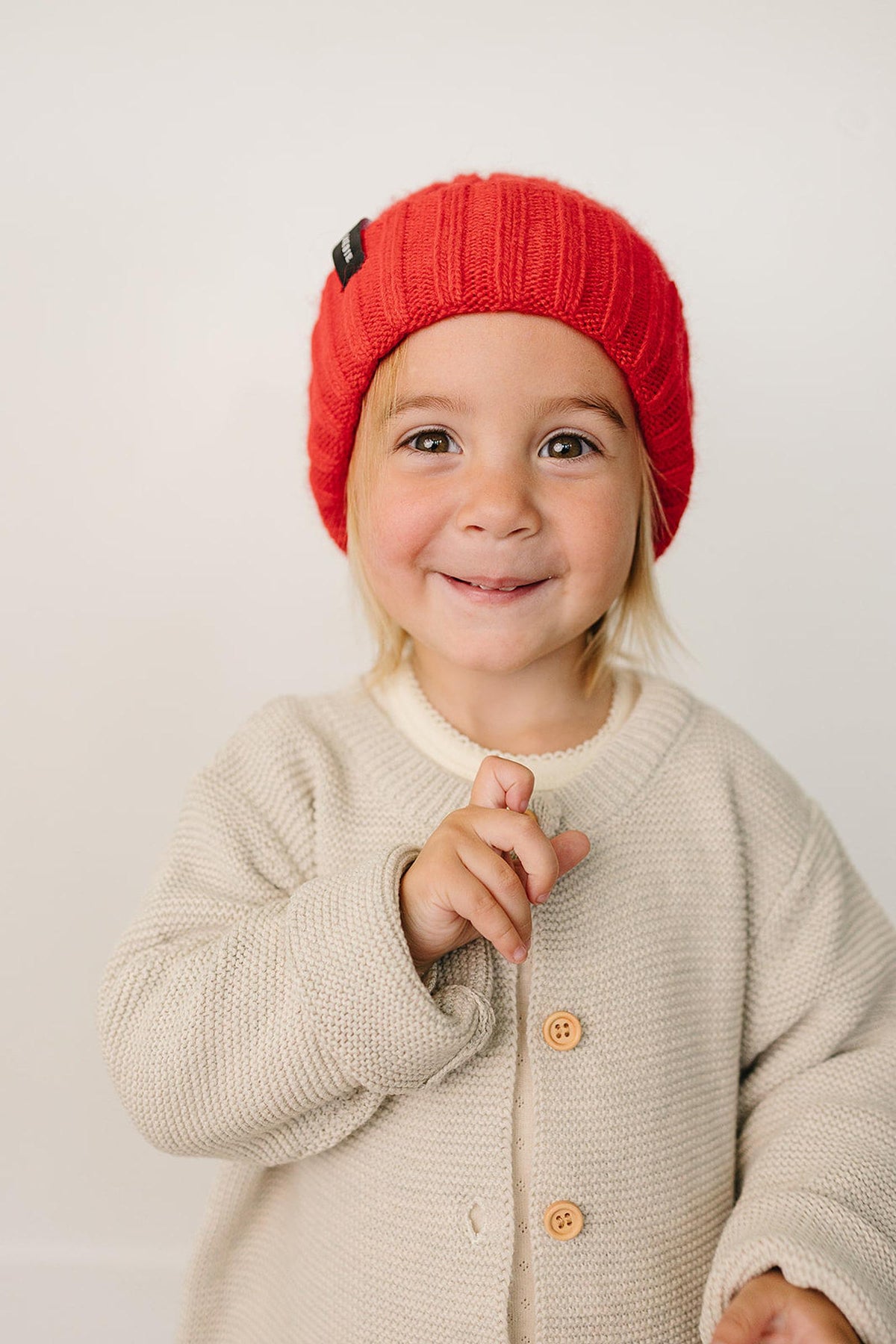 A child wearing a bright red chunky knit beanie smiles while holding up a finger, dressed in a cozy beige.