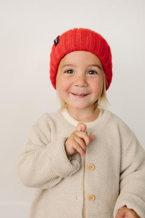 A child wearing a bright red chunky knit beanie smiles while holding up a finger, dressed in a cozy beige.