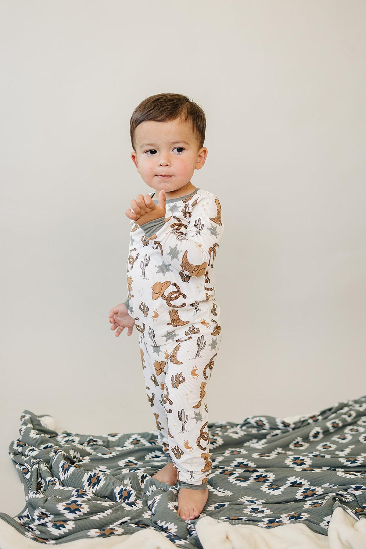A toddler wearing a cowboy-themed pajama set stands on a patterned blanket, featuring cacti and cowboy hats.