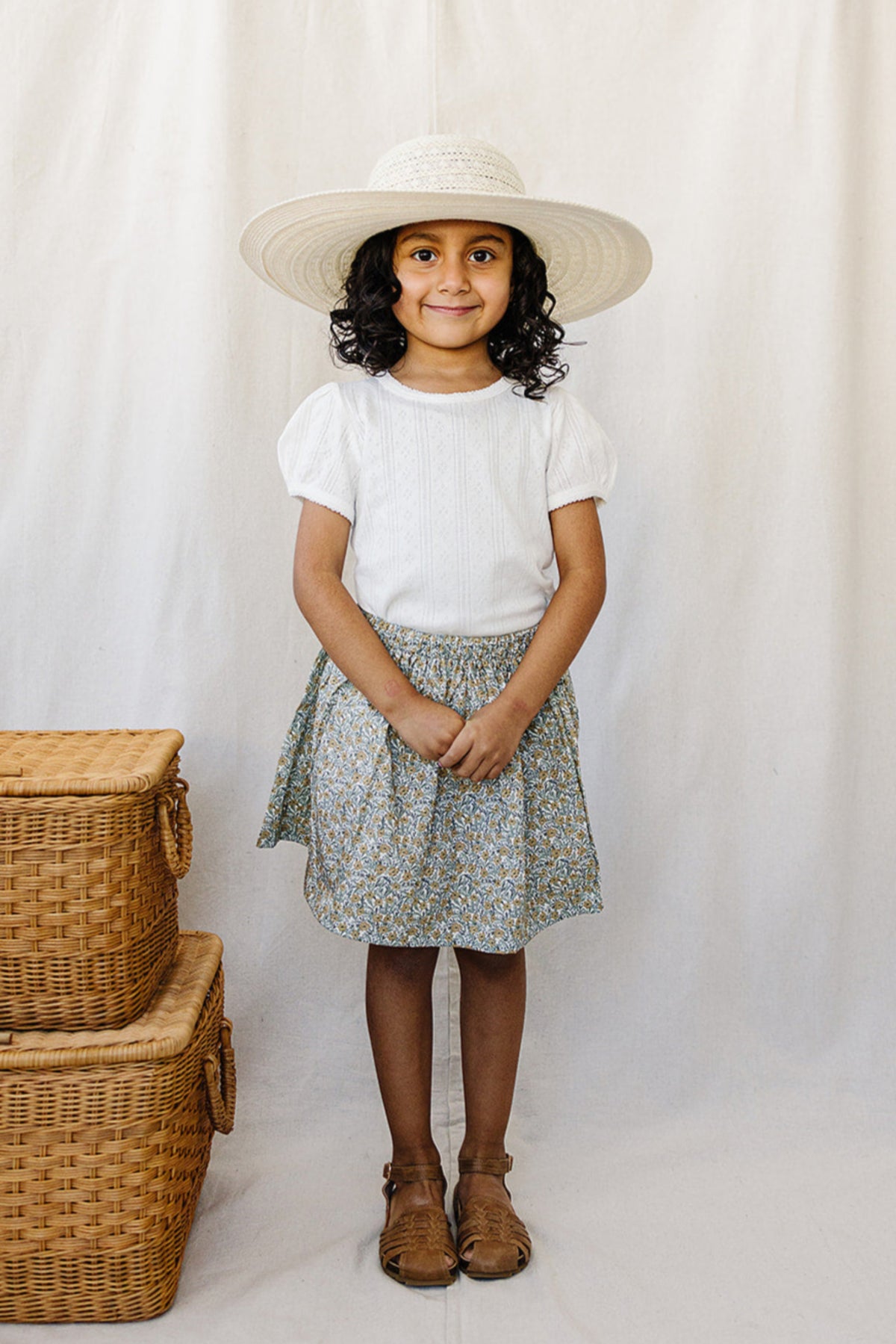 A young girl in a white pointelle t-shirt and floral skirt, wearing a wide-brimmed hat, stands smiling.