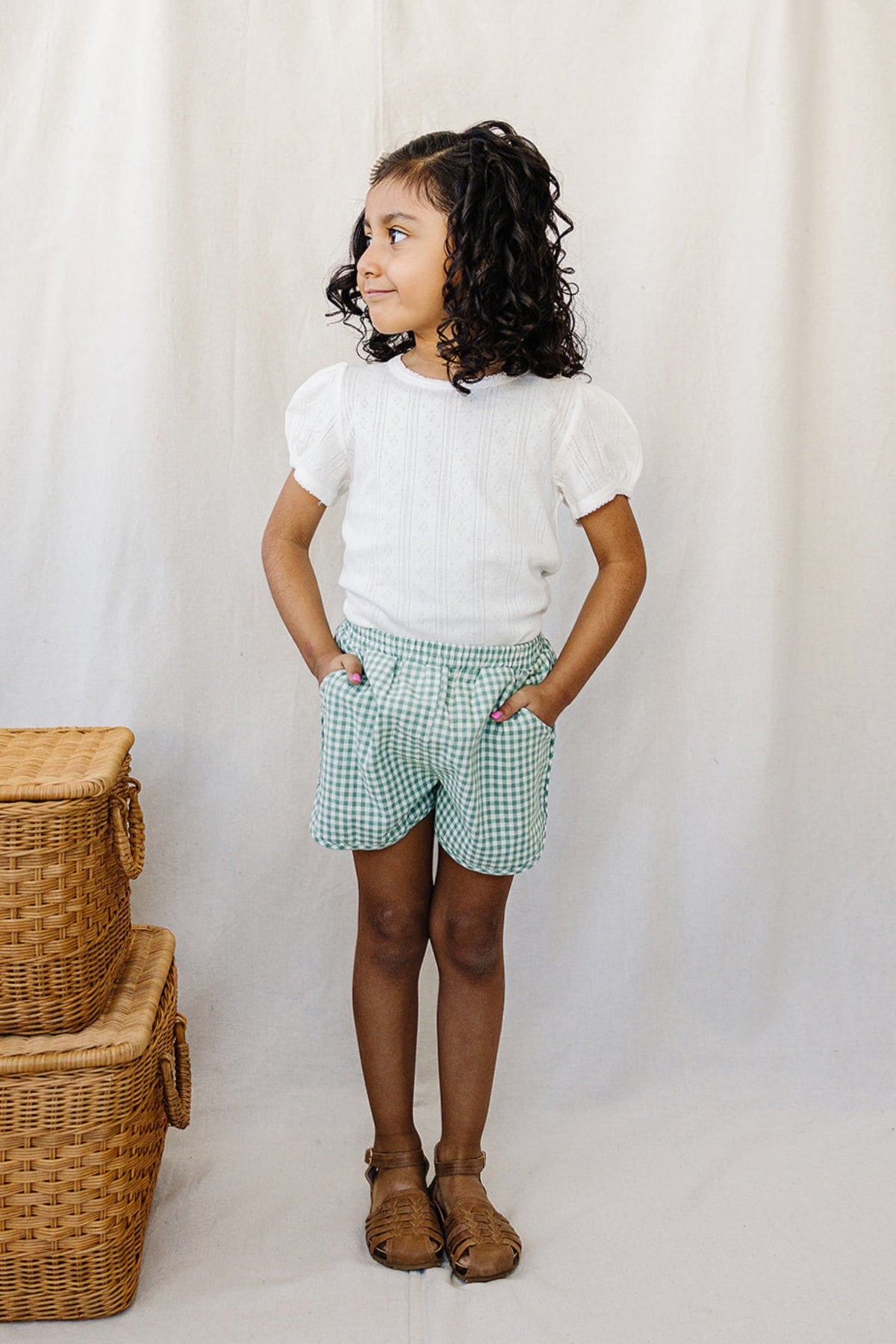 Young girl wearing a white pointelle t-shirt and green checkered shorts, standing beside wicker baskets on a beige background.