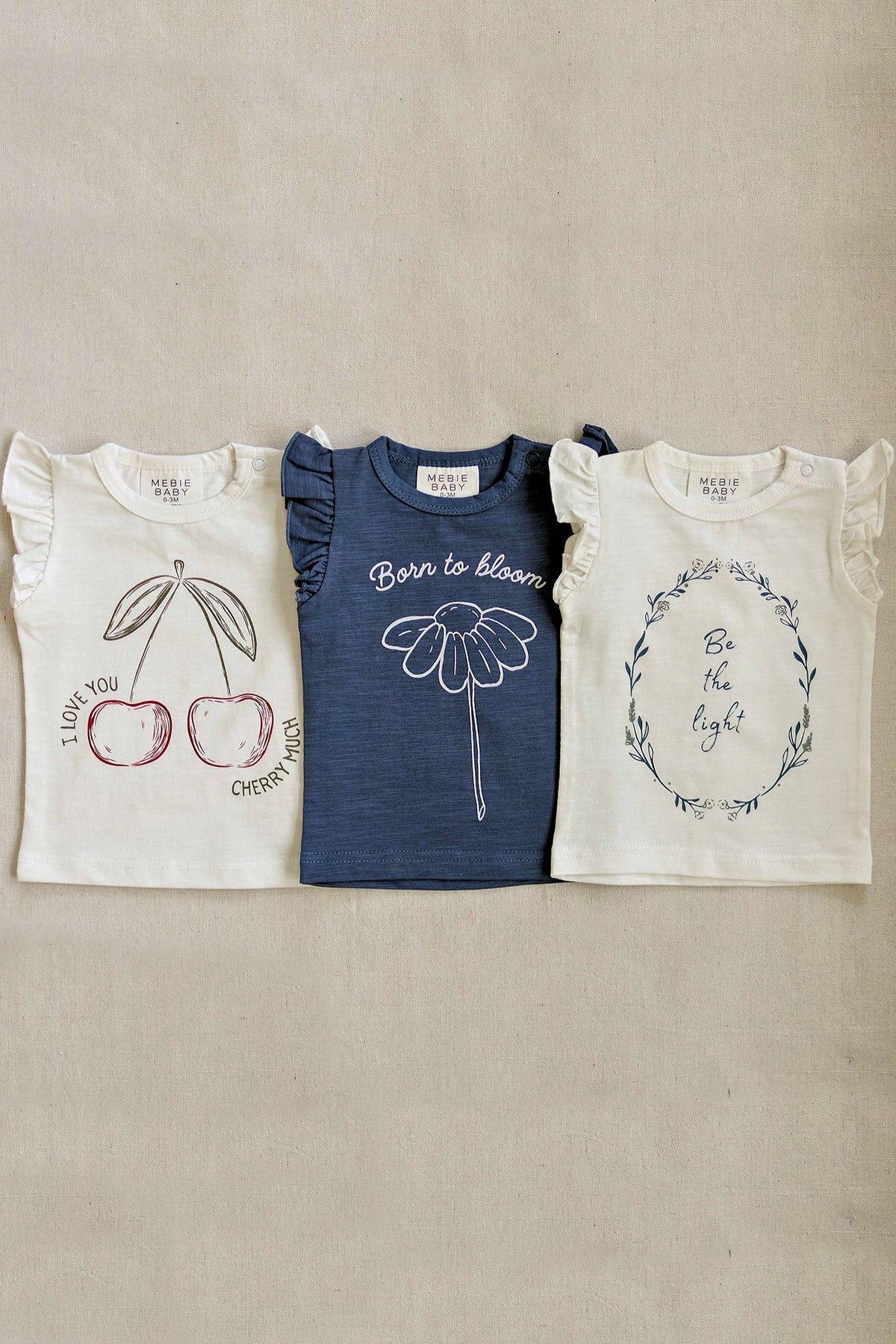 Three Mebie Baby t-shirts with ruffle sleeves feature cherry, flower, and floral wreath graphics on neutral tones.