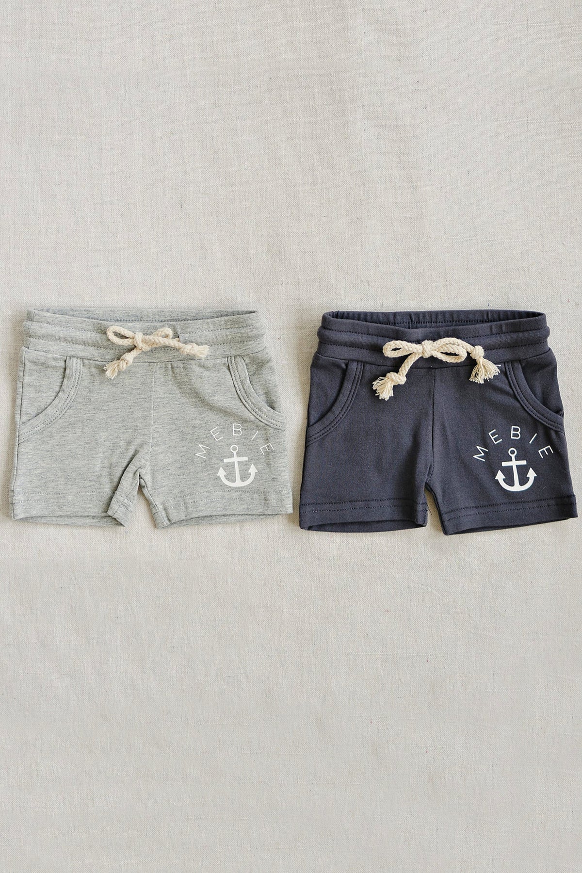 Two pairs of cotton shorts, one gray and one slate, with drawstrings and anchor logos on a beige background.