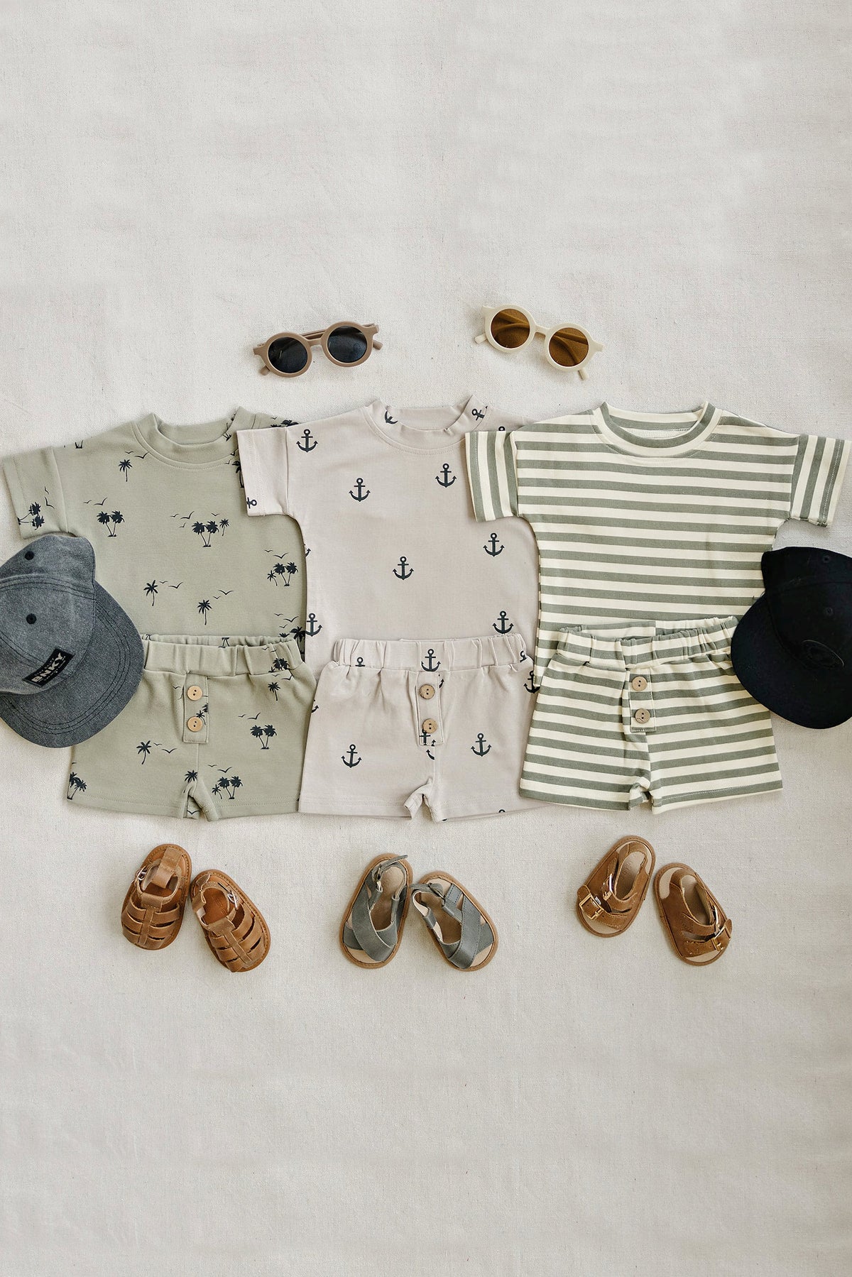 Three stylish baby and toddler outfits featuring short sleeve tops and elastic shorts in various colors, arranged with sunglasses and shoes on a neutral background. Perfect for warm weather adventures.