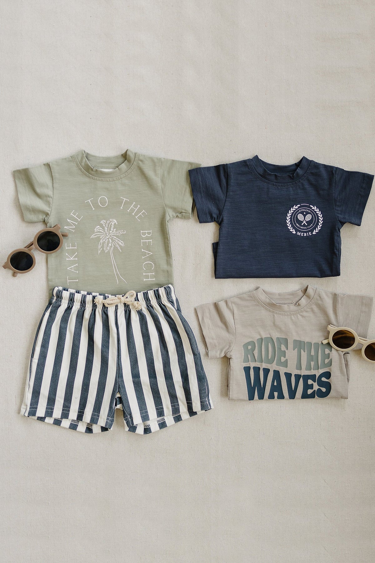 Three childrens T-shirts with playful designs and striped shorts are displayed on a neutral background with sunglasses.