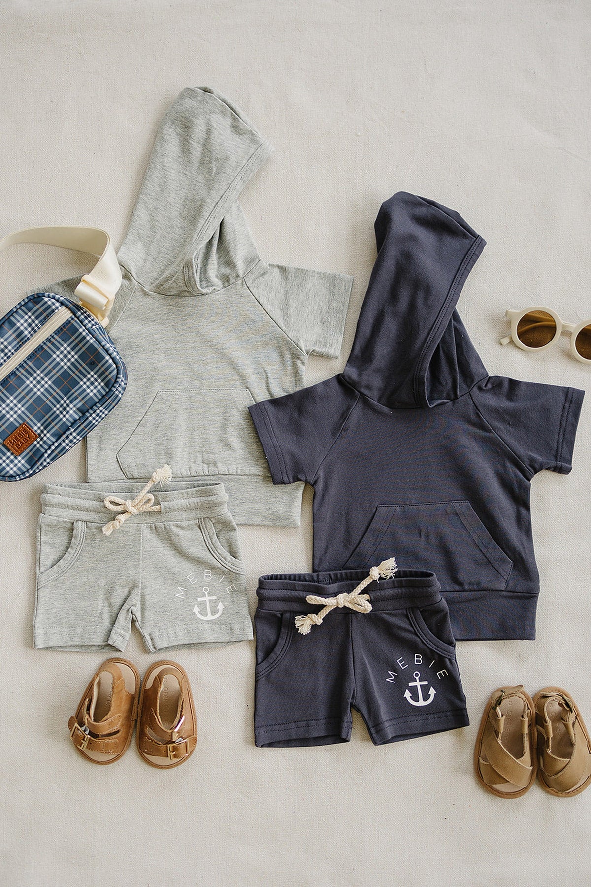 A heather grey hooded t-shirt and navy hoodie are displayed with matching shorts, alongside brown sandals and a plaid bag.