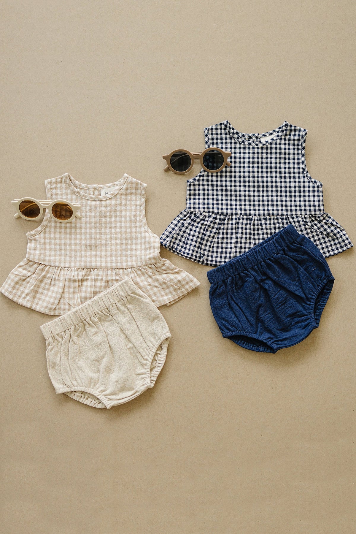 Two baby gingham peplum sets with matching bloomers, one in beige and one in navy, alongside sunglasses.