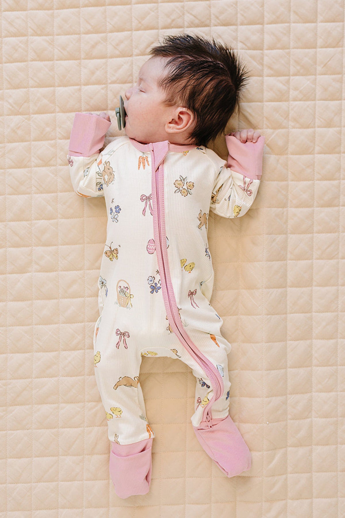 A sleeping baby girl lies on a quilted surface, wearing a soft, ribbed bamboo zipper pajama in a light cream color with pastel pink cuffs and whimsical spring-themed prints.