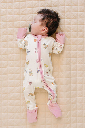 A sleeping baby girl lies on a quilted surface, wearing a soft, ribbed bamboo zipper pajama in a light cream color with pastel pink cuffs and whimsical spring-themed prints.