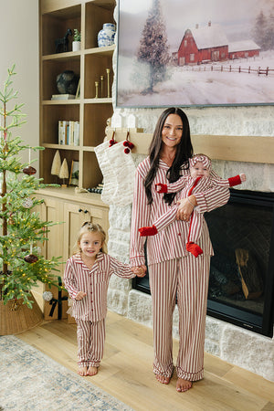 A woman holds a baby while standing next to a young child, both dressed in matching red striped pajamas. They are in a cozy living room decorated for Christmas.