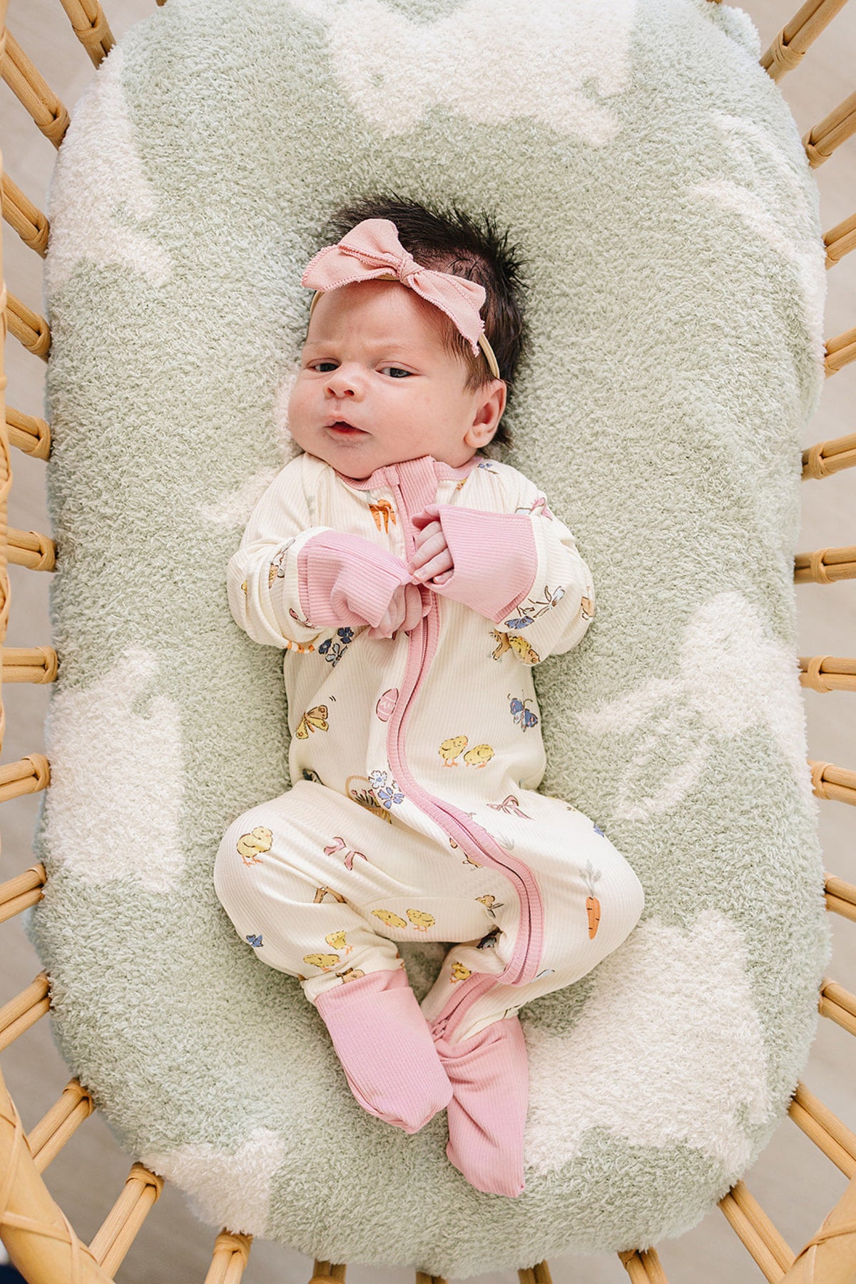 A newborn lies in a cozy bassinet, wearing a cream-colored bamboo zipper pajama adorned with playful animal prints and pink trim, complemented by a matching pink headband.