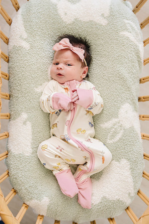A newborn lies in a cozy bassinet, wearing a cream-colored bamboo zipper pajama adorned with playful animal prints and pink trim, complemented by a matching pink headband.