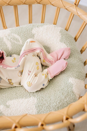 A baby girl in a cozy, ribbed bamboo pajama features a pastel print of bunnies and flowers, resting comfortably in a soft, cushioned basket.