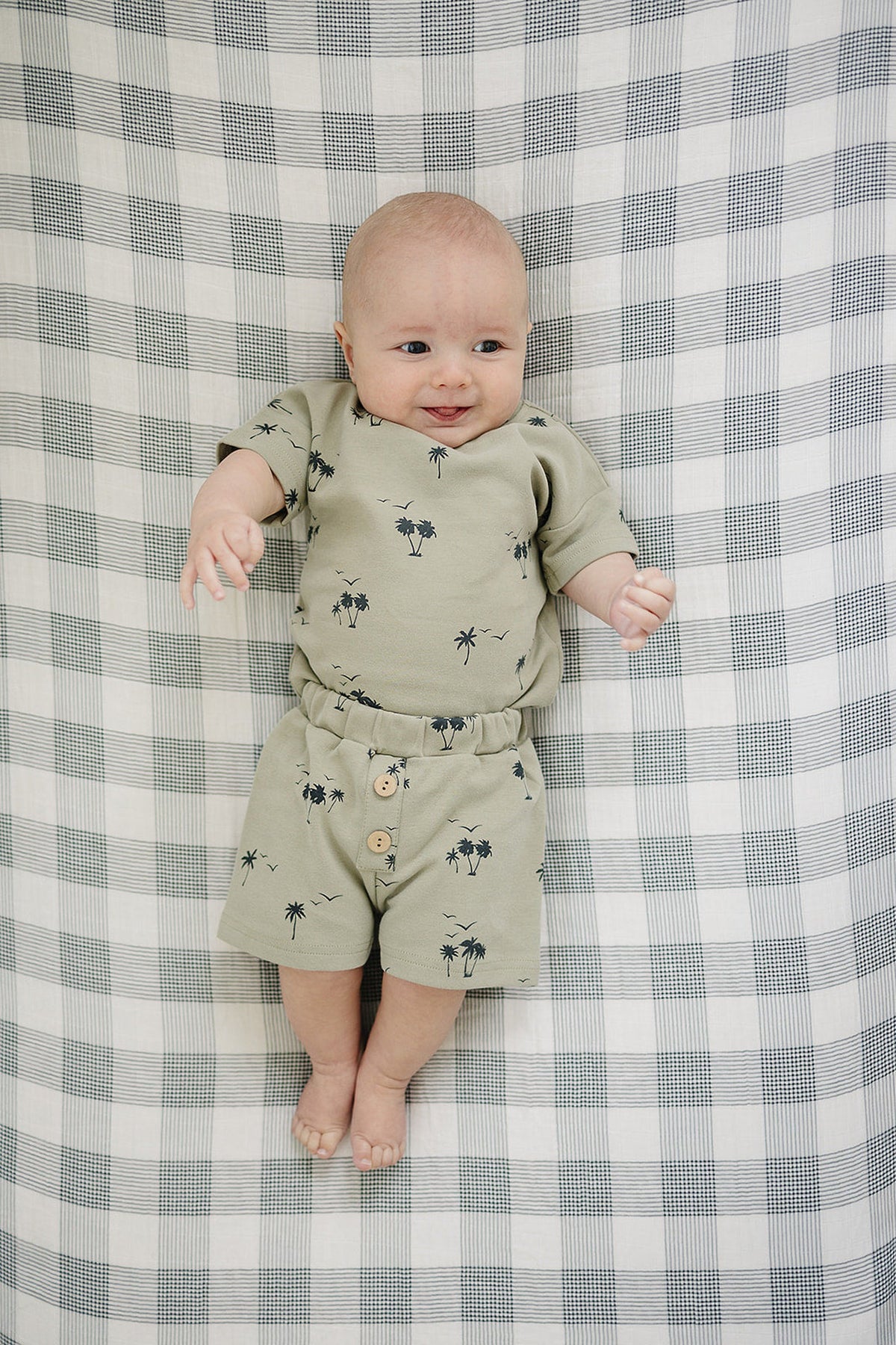 A happy baby lays on a checkered blanket, wearing a light green cotton outfit featuring palm tree prints and button details. The set includes a short-sleeve top and matching shorts.