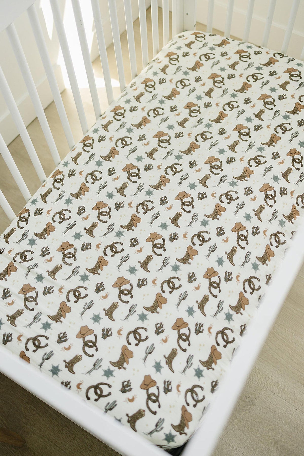 White crib sheet with cowboy-themed patterns, including boots and hats, in neutral colors, set in a white crib.