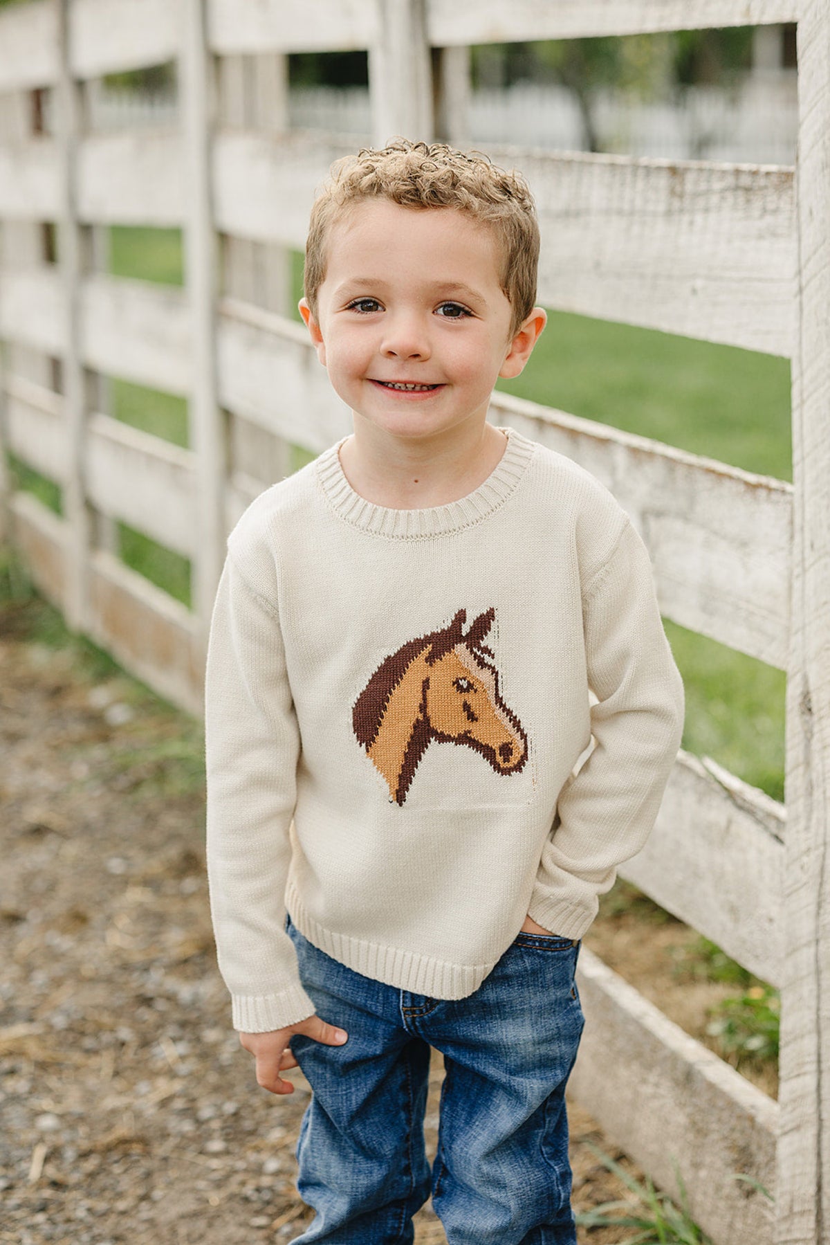 A smiling young boy stands by a wooden fence, wearing a cream-colored knit sweater featuring a horse design. His blue jeans complement the cozy, classic look of the outfit.