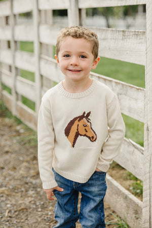 A smiling young boy stands by a wooden fence, wearing a cream-colored knit sweater featuring a horse design. His blue jeans complement the cozy, classic look of the outfit.