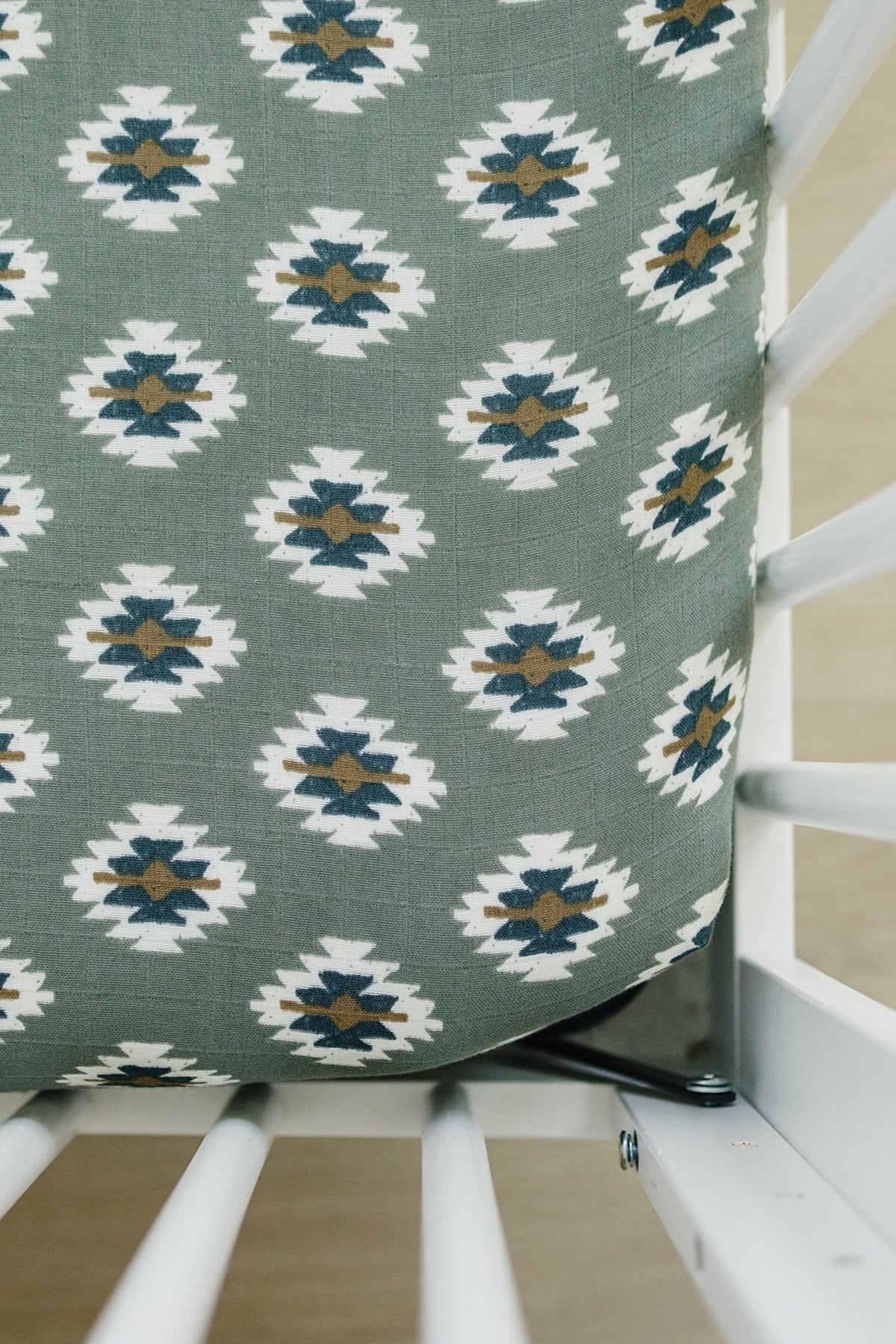 Gray crib sheet with white, brown, and navy Aztec pattern in a white crib. Made from soft muslin fabric.