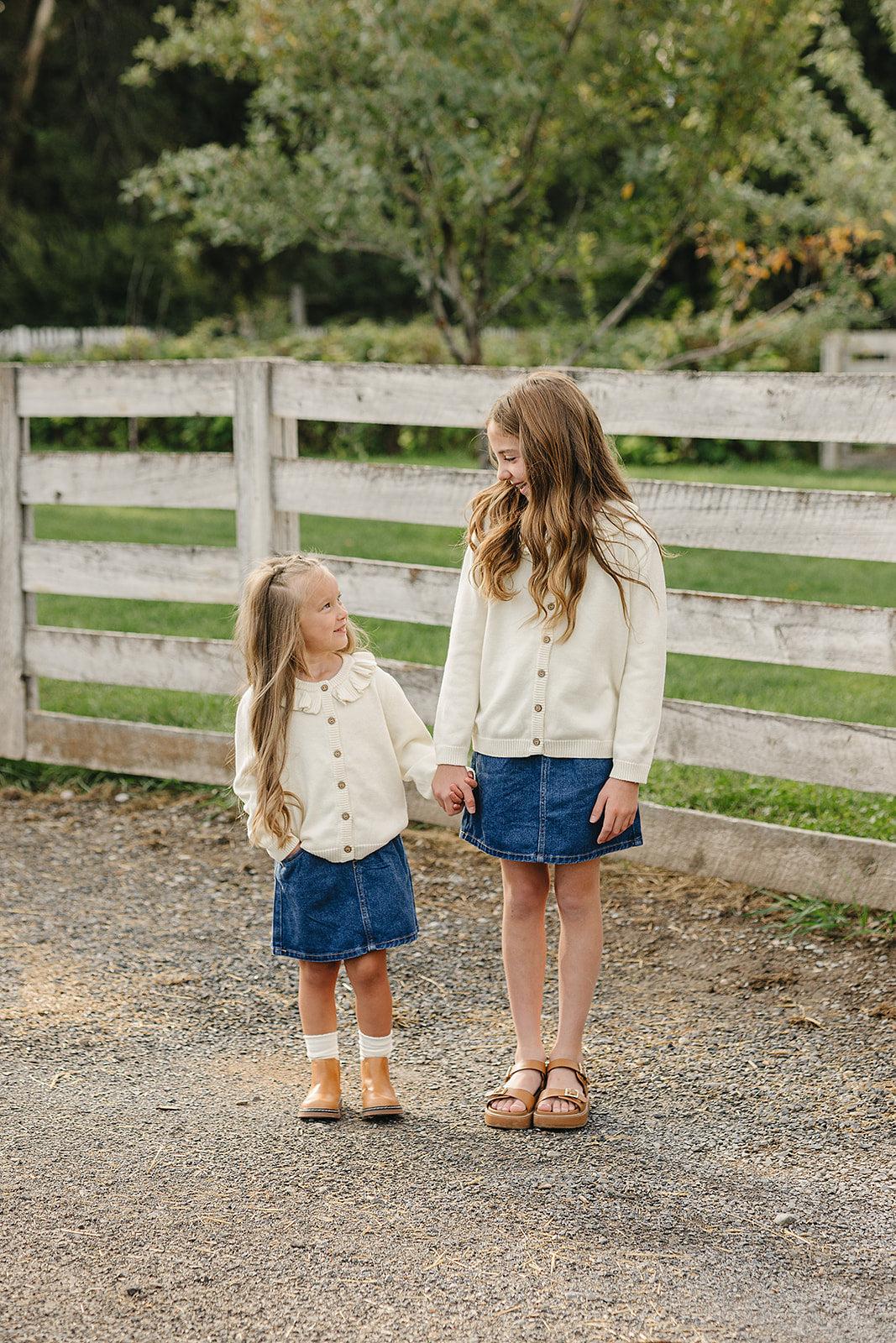 Two girls stand outdoors near a wooden fence, wearing matching cream ruffle knit cardigans and denim skirts.