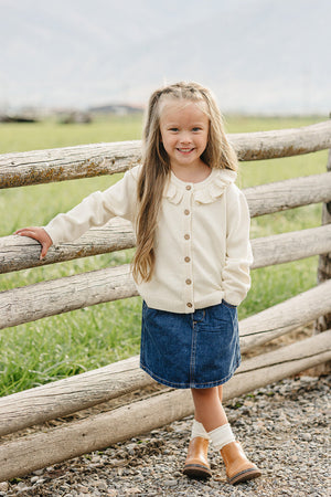 A young girl stands by a wooden fence, wearing a cream ruffle knit cardigan with wooden buttons and a denim skirt, showcasing a charming outdoor setting with green grass in.