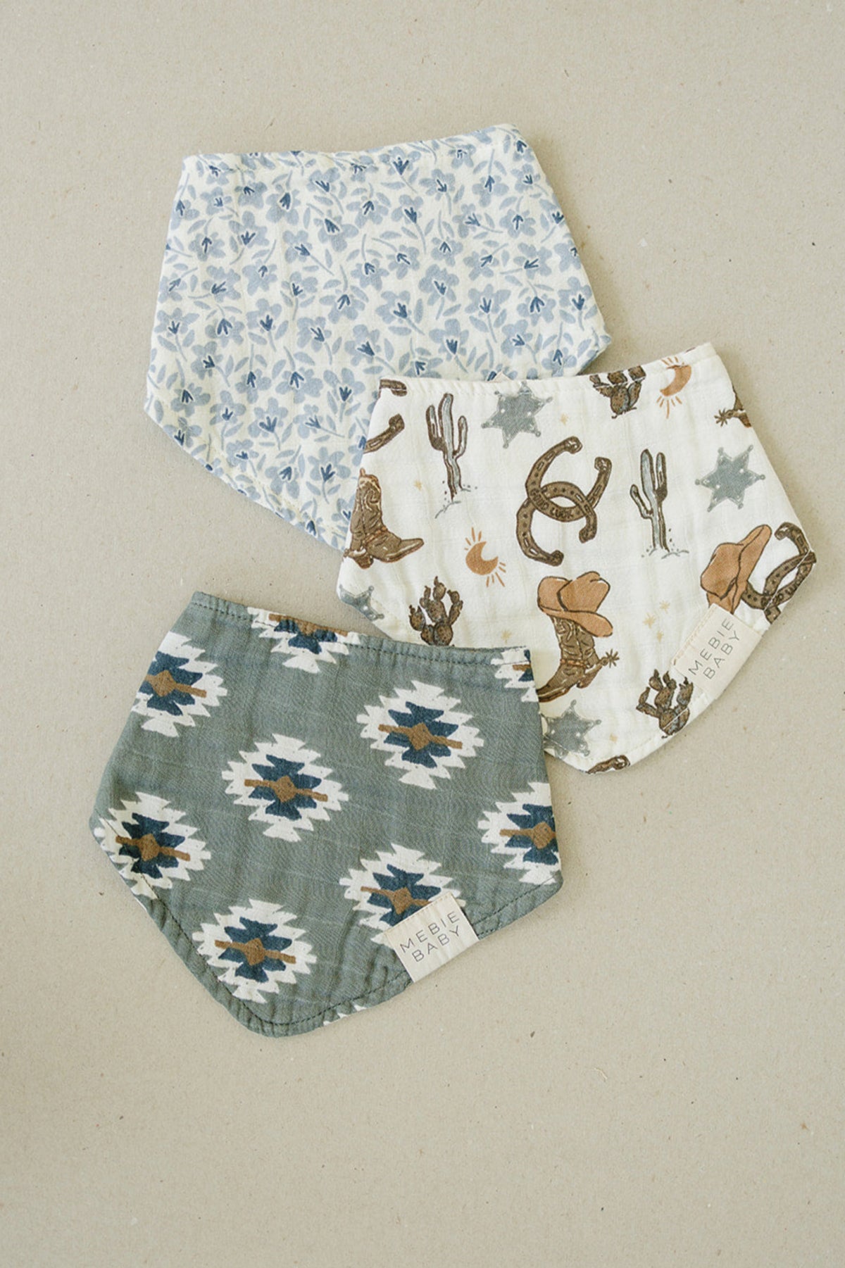 Three patterned muslin baby bibs are laid on a neutral background, featuring floral, cowboy, and geometric designs.