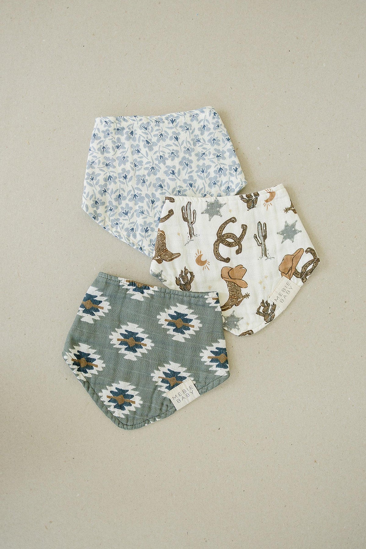 Three Mebie Baby muslin bibs with blue floral, Southwest Aztec, and cowboy-themed prints on a neutral background.