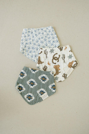 Three Mebie Baby muslin bibs with blue floral, Southwest Aztec, and cowboy-themed prints on a neutral background.