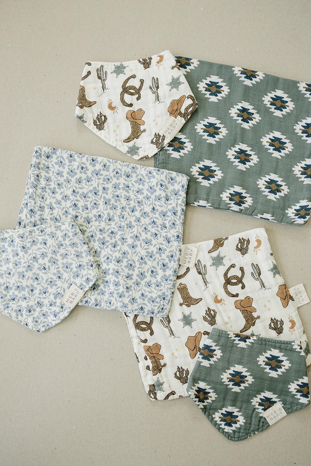 Assorted muslin bibs with cowboy, floral, and geometric patterns are arranged on a neutral background.