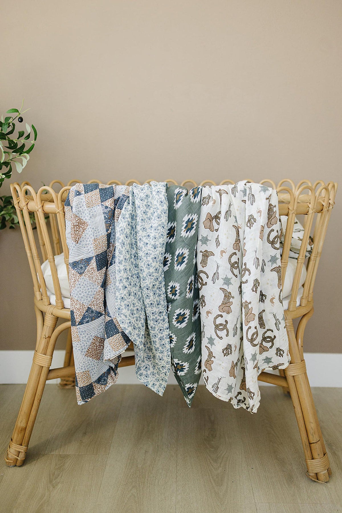 Four colorful muslin swaddle blankets with various patterns draped over a bamboo crib in a neutral room.