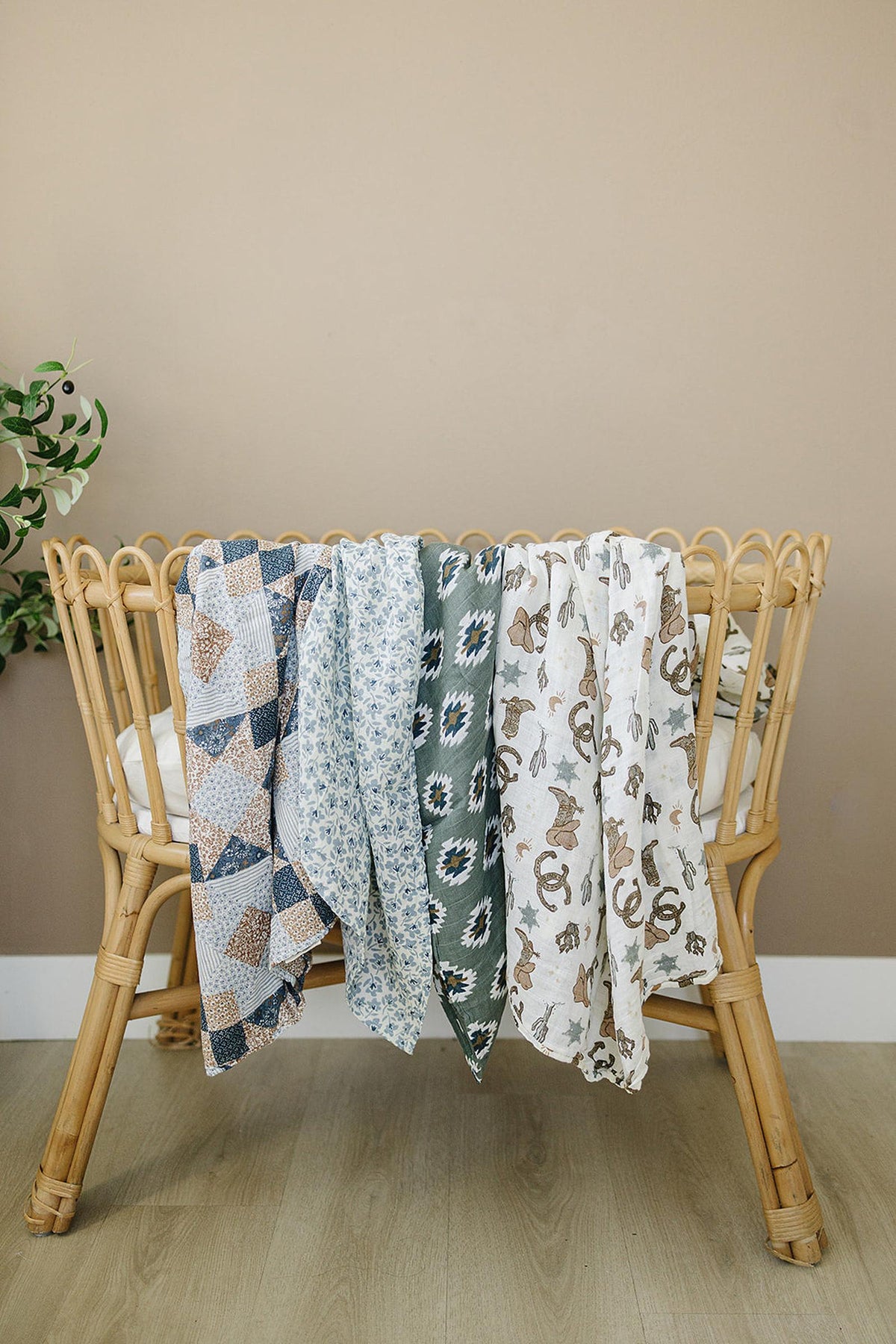 Rattan crib with various patterned muslin swaddle blankets draped over the side, including floral and cowboy designs.